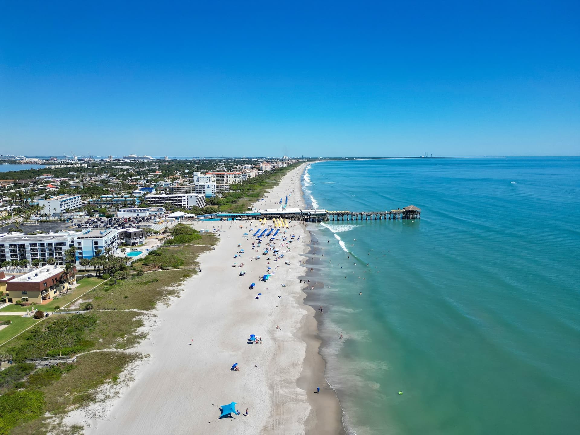 Aerial view of Cocoa Beach Florida coastline with turquoise ocean, white sandy beach, colorful umbrellas, and the iconic fishing pier